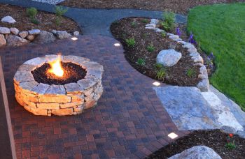 Circular stone fire pit burning on a brick patio in a landscaped backyard at dusk